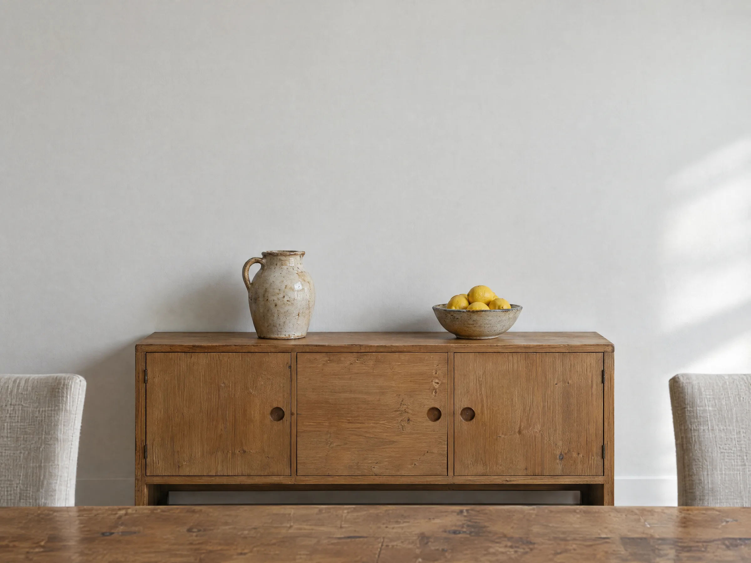 A low oak sideboard with a stoneware jug and a bowl of lemons against a chalk white dining-room wall