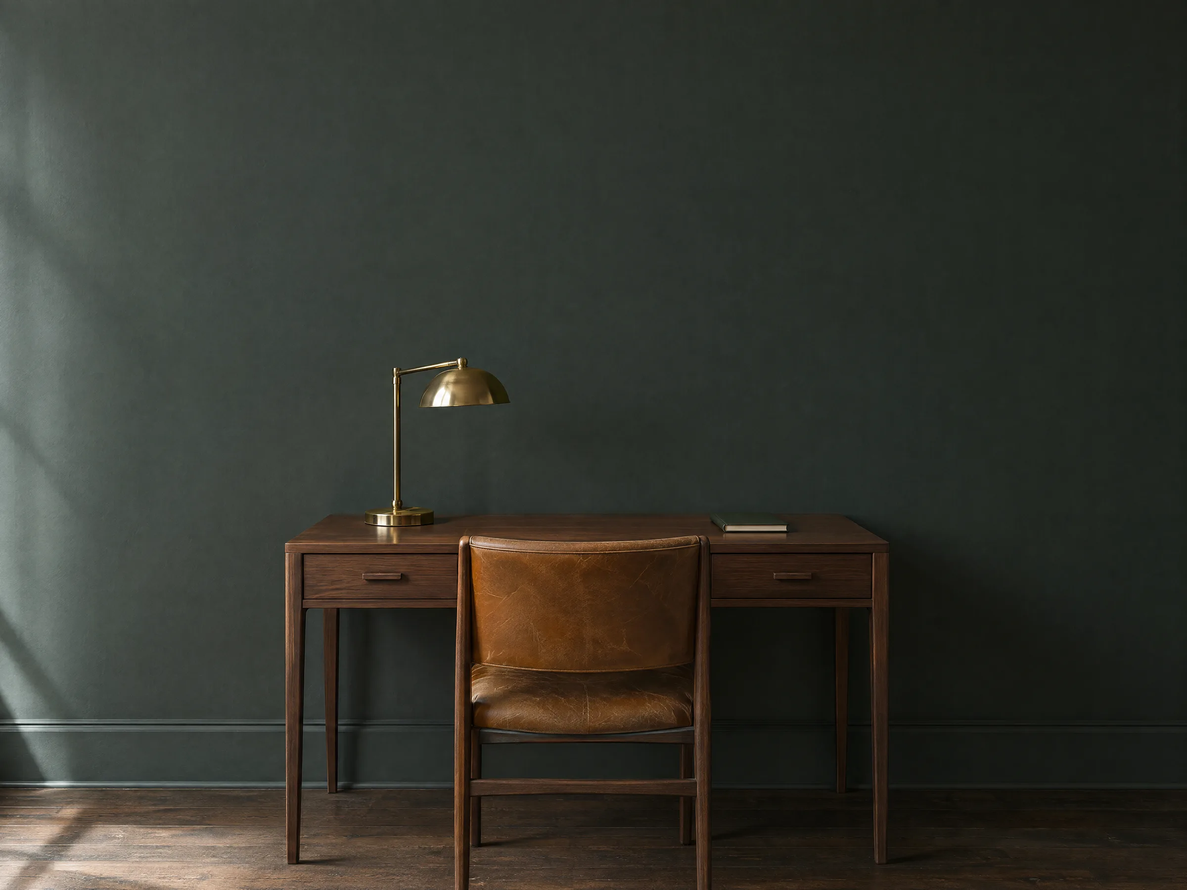 A walnut writing desk with a brass lamp and a tan leather chair against a deep green-grey wall